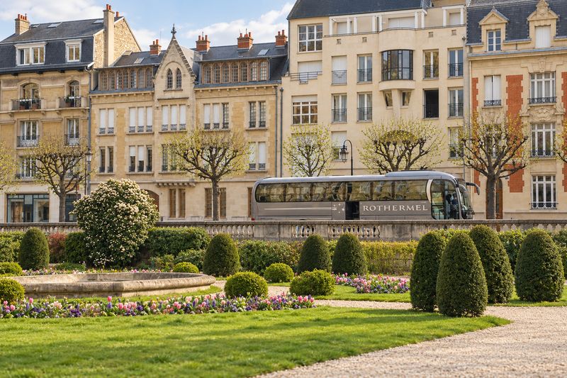 Grauer Reisebus von Rothermel Reisen an einer Parkanlage vor historischen Stadthäusern